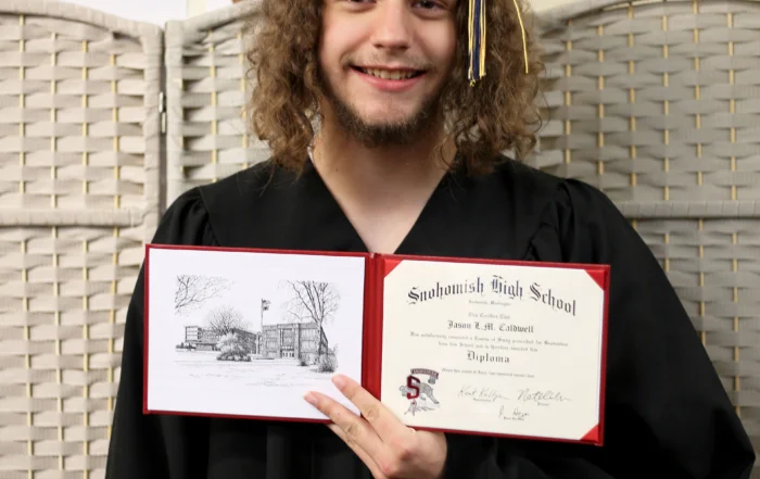 A man in a graduation robe and hat smiles and holds up a diploma.