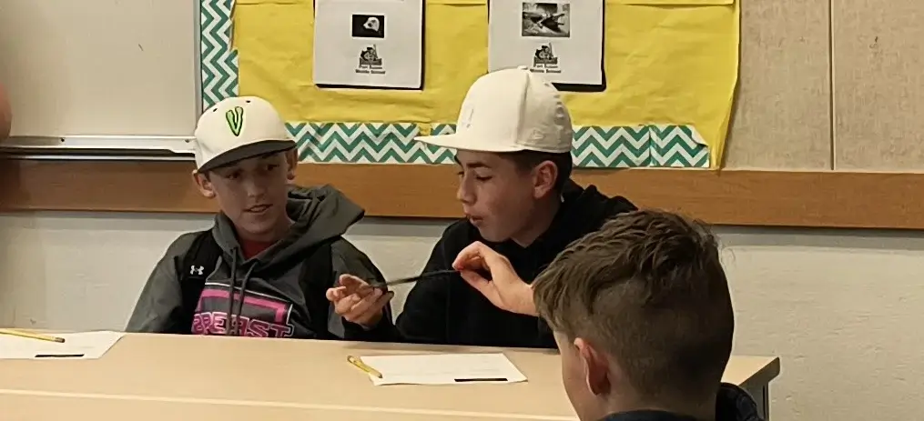Two students in hard hats hold pieces of machinery while seated at a desk.