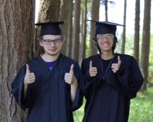 IMG_3363 Two boys stand in graduation caps and gowns, giving thumbs up signs in the trees.